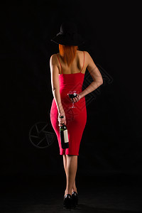 穿着亮红礼服的感女孩戴着黑色宽胸帽子拿着酒杯和瓶站在黑暗背景上图片