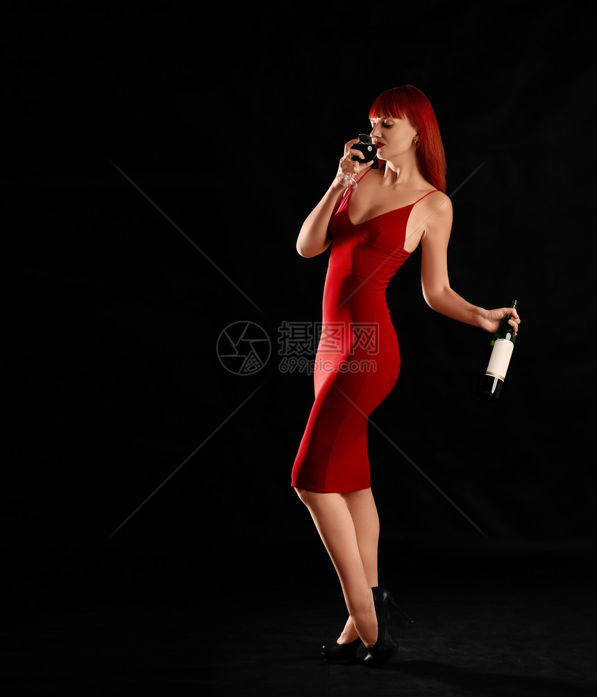 穿着红礼服的感发女孩站在黑暗背景上一杯酒和瓶红发和葡萄图片