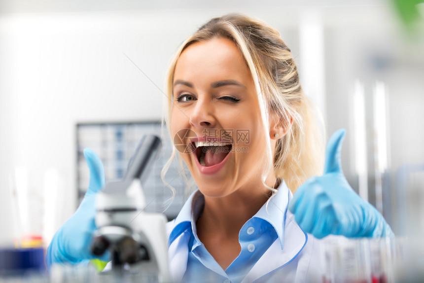 在科学化实验室露出拇指的快乐年轻迷人笑女科学家肖像图片
