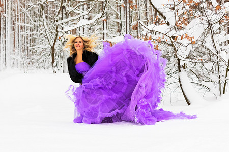 穿着长紫色裙子的金发年轻美女在雪的冬季森林或户外的木里背景图片