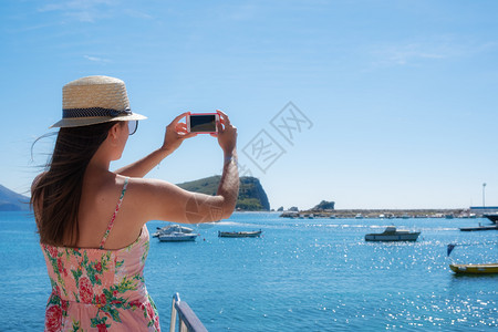 戴帽子的年轻女旅行青用智能手机相拍摄夏日阳光明媚的海景照片图片