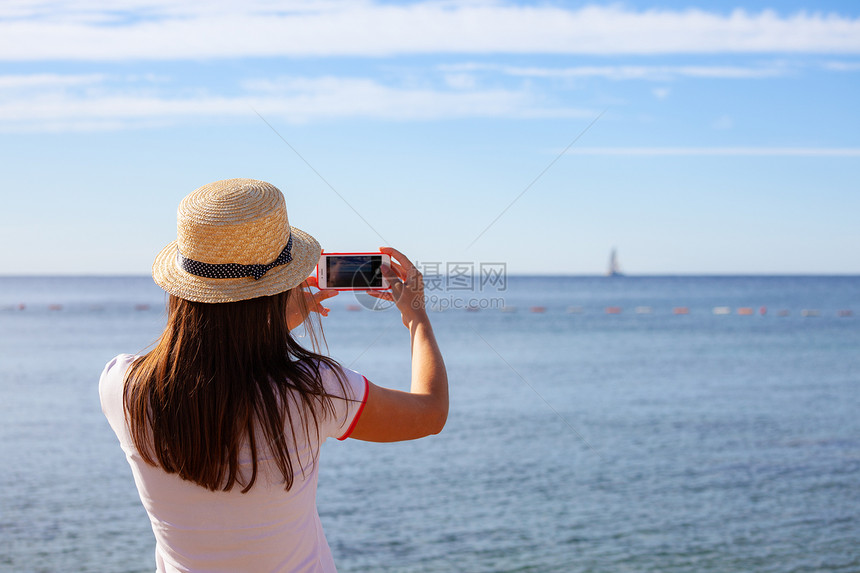 戴帽子的年轻女旅行青用智能手机相拍摄夏日阳光明媚的海景照片图片