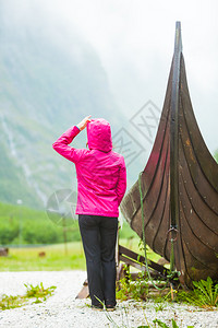 在挪威自然界的旧木型维京船附近站立的女旅游者看着迷雾山雨日旅游和概念在挪威自然界的旧木型维京船附近站立的旅游者背景图片
