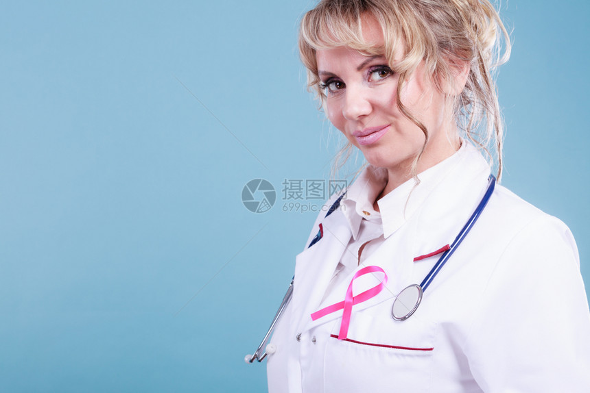 乳腺癌医疗帮助治概念穿白色围裙蓝底带粉色丝的女金发医生穿粉色癌症丝带的医生图片