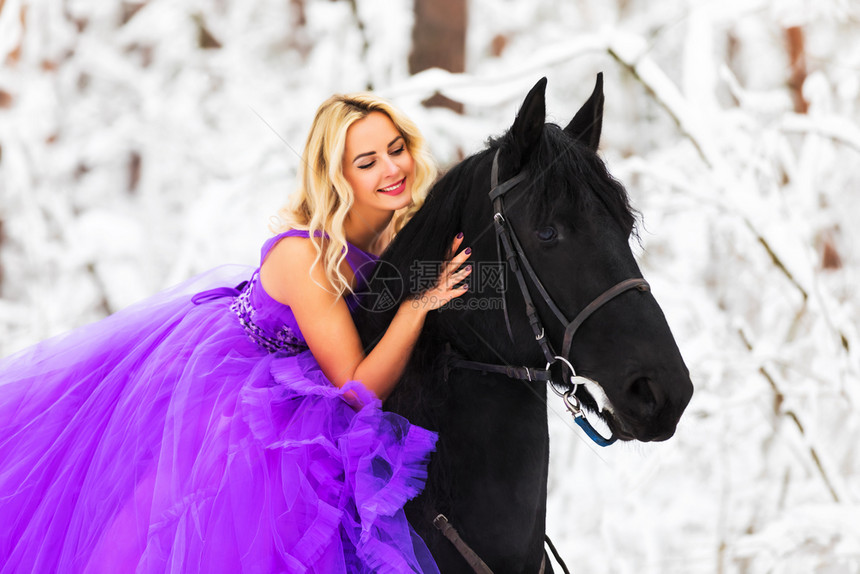 身穿长的粉红裙子在雪冬林骑着黑马的年轻金发美女图片