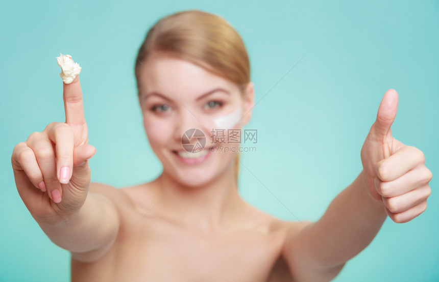 美容治疗女在脸上使用润湿奶油护肤产品拍出绿色蓝底的拇指牌工作室女在皮肤上涂奶油图片