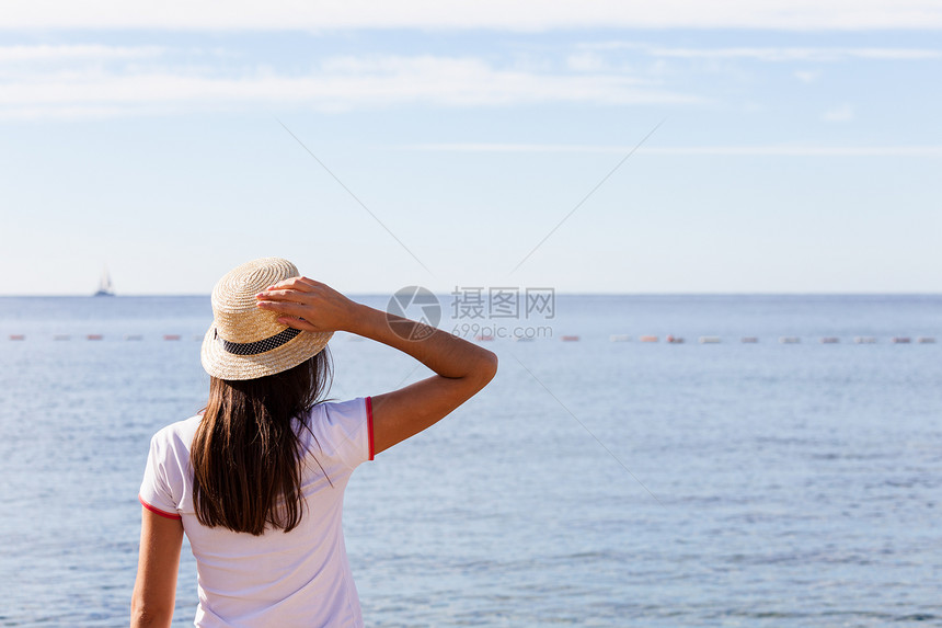 美丽的女孩看着海戴帽子的年轻女孩看着平静的海蓝天后视风戴着帽子的年轻女孩看着海图片