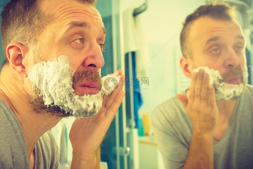 男人脸上涂着剃须泡沫奶油站在浴室里看着镜子准备刮胡图片