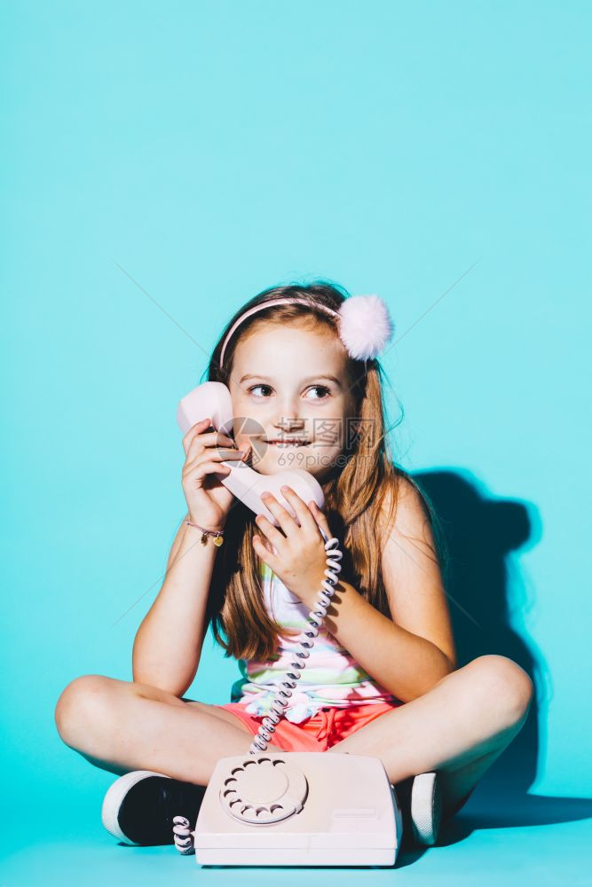 小女孩使用粉色模拟电话坐在蓝背景上电话交谈小女孩使用粉色模拟电话图片