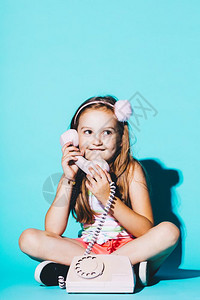 小女孩使用粉色模拟电话坐在蓝背景上电话交谈小女孩使用粉色模拟电话图片