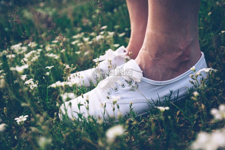 穿着白色运动鞋的女子站在春草地上带着鲜花站脚贴近有活力穿着运动鞋的女子站在草地上拿着鲜花站图片