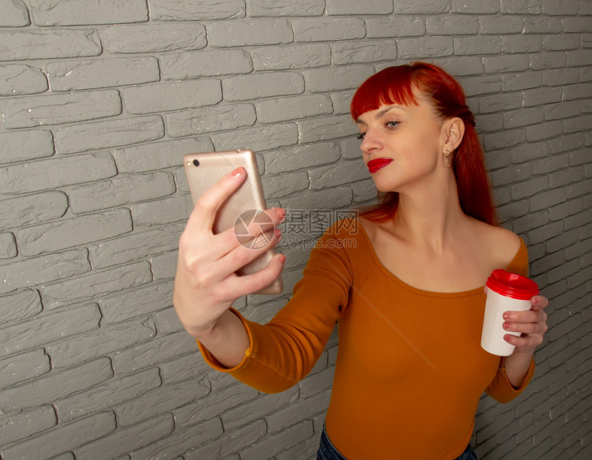 年轻红头发女孩带着一杯咖啡用她的智能手机在灰砖墙上做自拍女孩图片