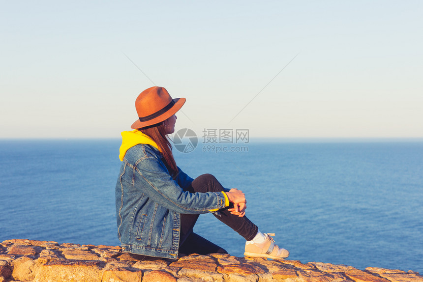 穿着牛仔裤夹克戴帽子坐着看蓝色的海洋和天空图片