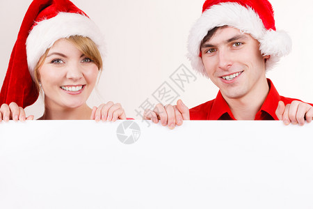 白色招牌素材一对持有横幅标志的夫妇带有文本复制空间在的广告牌边上偷看快乐的女孩和男在圣达克拉斯的帽子中圣诞广告复制空间的横幅板上夫妇圣诞背景