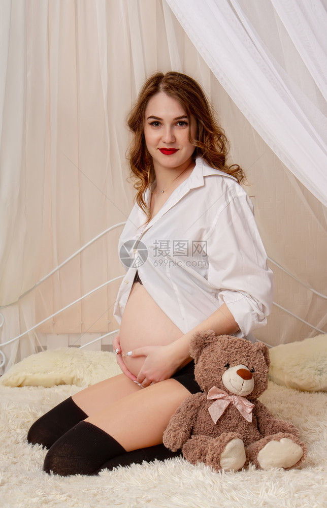 怀孕6个月时一美丽的年轻女孩坐在她丈夫的轻便床上穿衬衫摸她的肚子图片