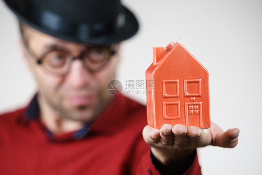 戴着小红房子模型的带滑稽帽子和眼镜的成人男子是房地产经纪人家庭所有权概念图片