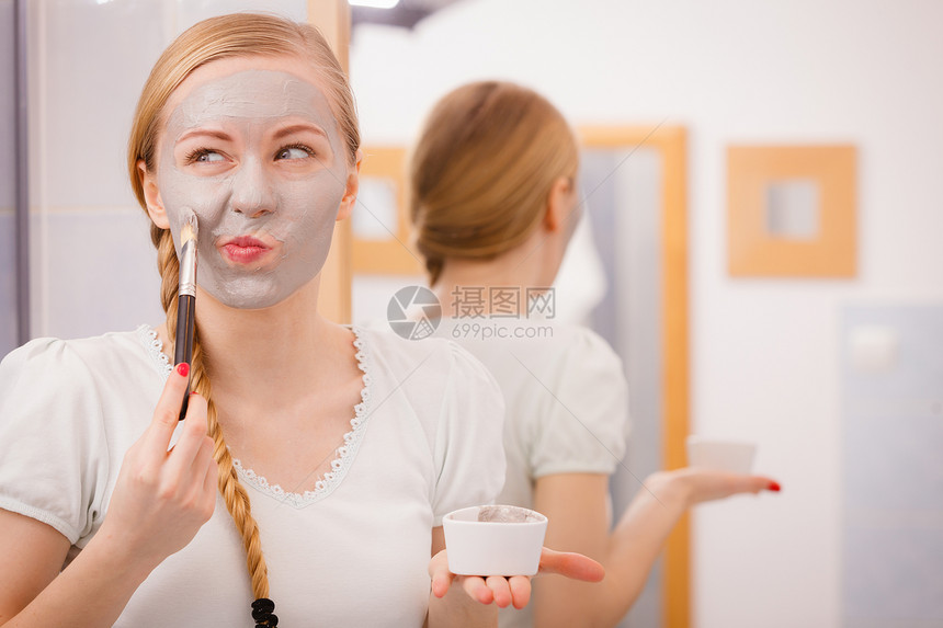 面部干燥皮肤和身体护理家用皮肤处庭概念的皮肤处理年轻女在脸上戴灰泥面罩用刷子做滑稽的脸孔快乐年轻女在脸上戴泥面罩快乐的年轻女在脸图片