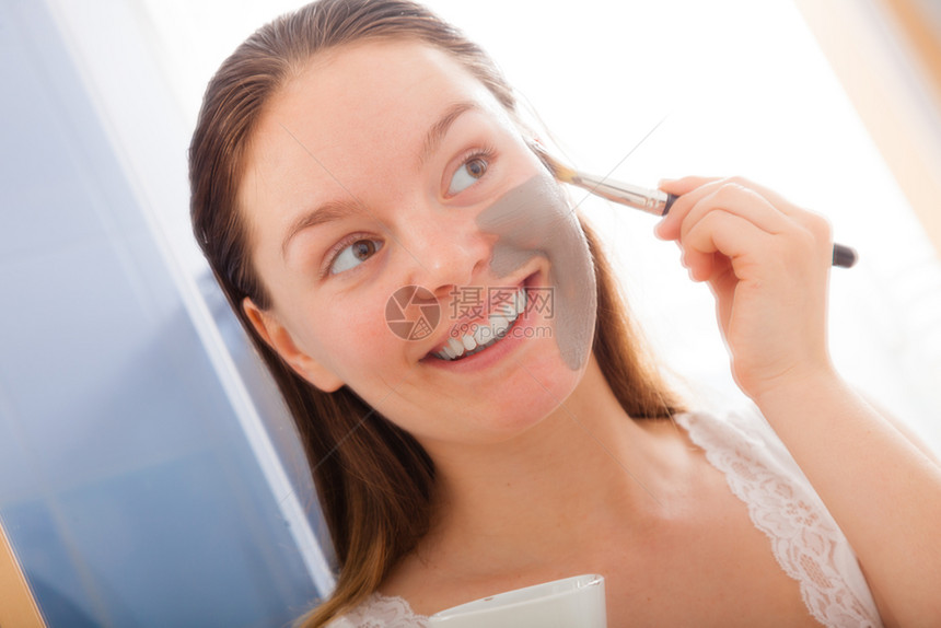 美容程序皮肤护理概念年轻妇女没有化妆品在洗手间用刷子将面部灰色泥粘土面具套在脸部上图片