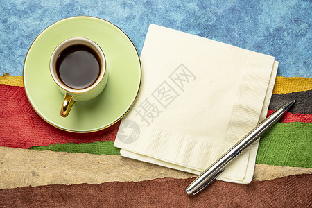 餐巾纸笔和一杯咖啡与用纸质的彩色手工造纸所创的抽象景观相对背景图片