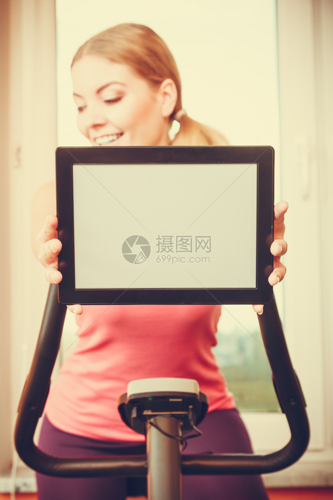 参加运动的年轻女从事运动的自行车固定式工作手持Pc平板电脑有空白的屏幕复制间在家对运动女生进行培训关于健身和减肥的广告概念图片