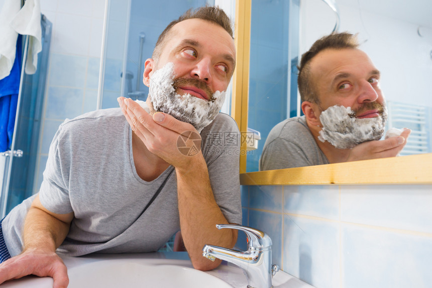 男人脸上涂着剃须泡沫奶油站在浴室里看着镜子准备刮胡图片