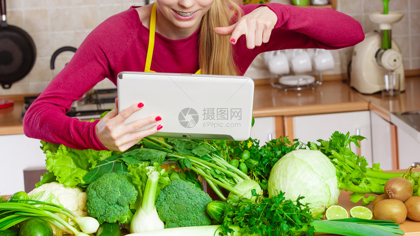 厨房的年轻女在餐桌上吃着许多绿色蔬菜拿着平板电脑思考烹饪的东西在互联网上寻找食谱图片