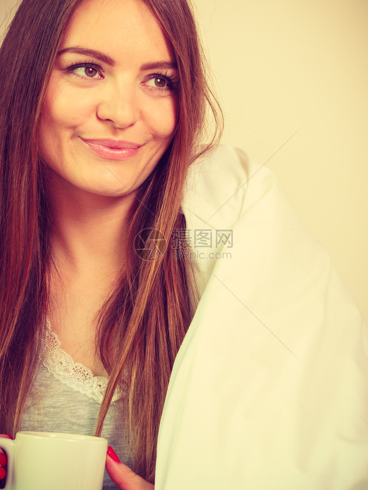 美丽的微笑年轻女子坐在室内的床上喝着热咖啡或茶图片