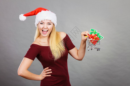 戴圣诞老人帮手帽子的快乐妇女拿着小圣诞树和里面的礼物购篮车背景图片