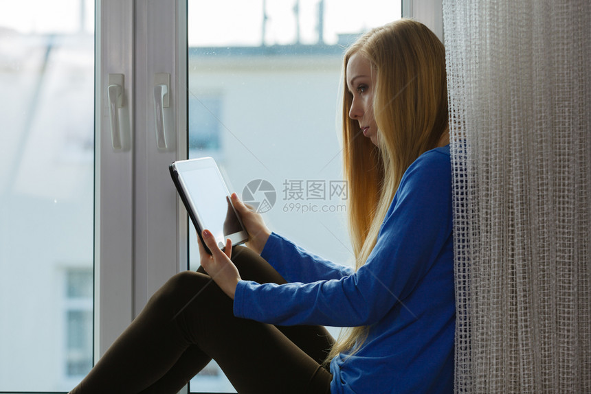 快乐的年轻少女在坐窗台上时使用平板在互联网上放松冲浪图片