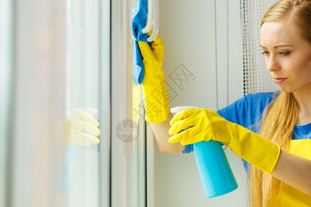 穿黄色手套的年轻女士用蓝色抹布和喷雾洗涤剂擦窗户春季清洁家务概念用洗涤剂擦窗户的女孩在家里用洗涤剂擦窗户背景图片