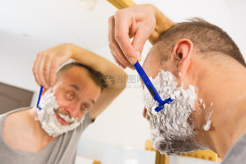 脸上有很多剃须泡沫的男人站在浴室里看着镜子刮胡图片