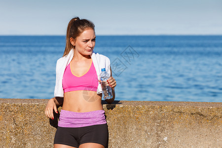 穿运动服的妇女休息用塑料瓶水补充饮用在海边户外运动锻炼后休息图片