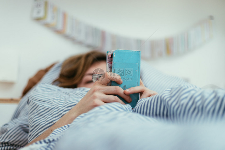 躺在床上的女孩正在使用她的智能手机在前台模糊的背景图片