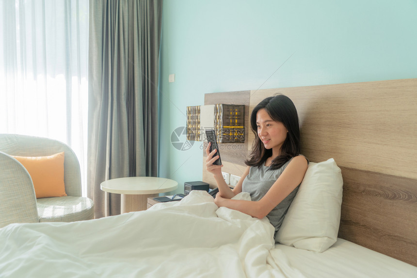 快乐的亚洲女人微笑使用手机和朋友在床上聊天现代卧室里图片
