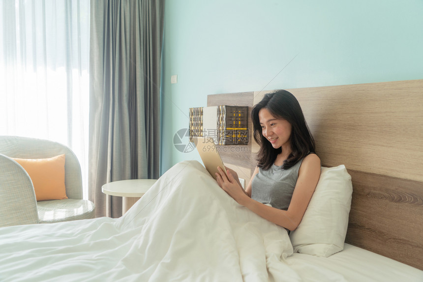 快乐的亚洲女人笑着用平板在床上现代卧室里早上带着白毯子图片