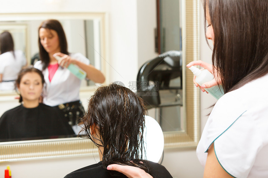 穿着黑斗篷的女士在美容院师用理发喷洒她的头图片