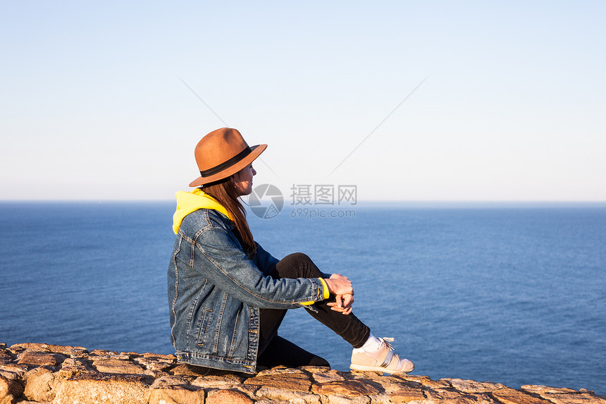 穿旅行服装和戴帽子的妇女坐着看蓝海和天空旅行概念照片坐着和看蓝海天空的妇女图片