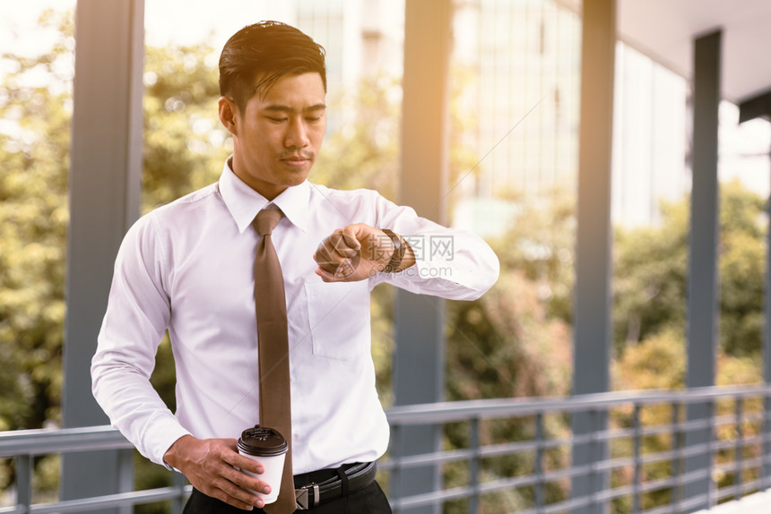 年轻亚裔男站着检查时间拿咖啡杯满怀信心地看着外面公司手表图片