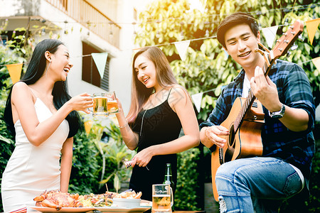 亚裔年轻女在家庭花园户外与玩吉他的人一起享受敬酒派对图片
