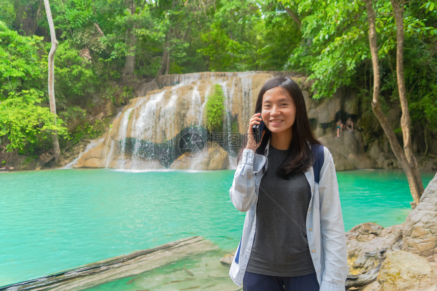 在泰公园旅行和度假期间快乐的亚洲女使用移动智能手机在热带森林的Erawan瀑布上聊天或致电社交媒体朋友图片