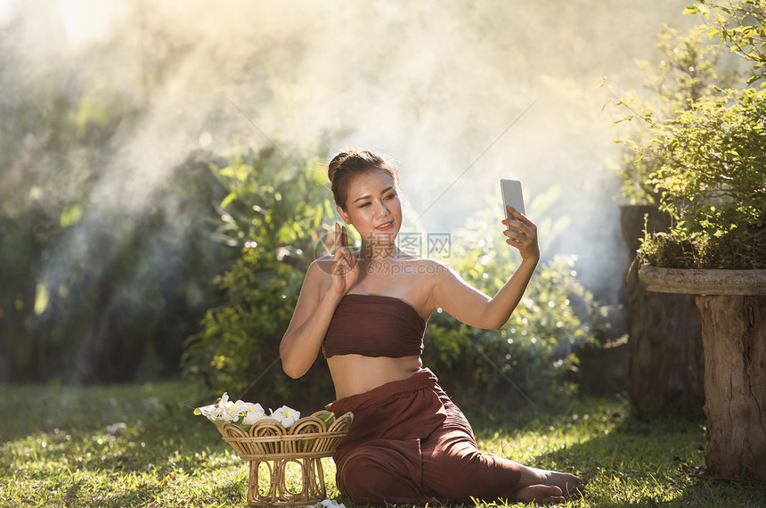 亚裔女微笑的年轻孩在花园里用智能手机拍自肖像泰国女在传统服装戏剧中拍泰式图片