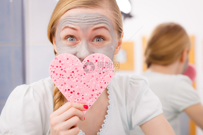面部干燥皮肤和身体护理家用皮肤处概念快乐的年轻女喜欢脸上有灰色的泥面罩将海绵放在心脏形状中灰色面罩的妇女则戴着心海绵图片