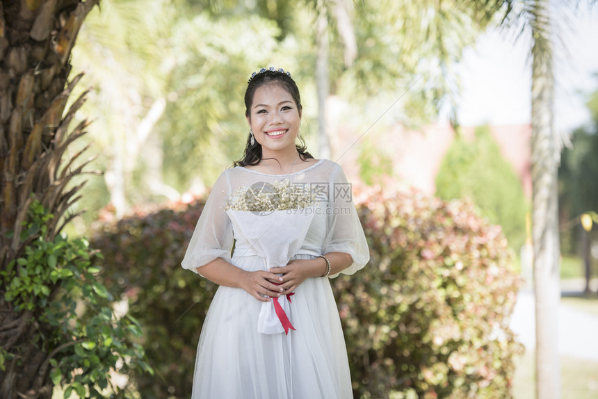 美丽的亚洲女新娘肖像在户外花园公中微笑并握着花束图片