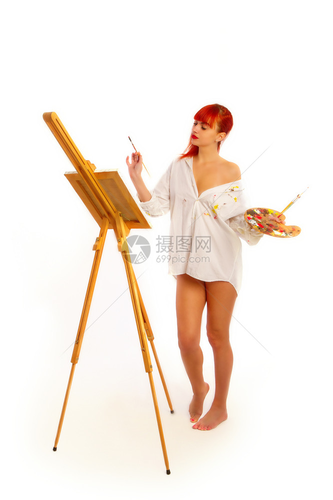 一个穿着涂漆烟雾衬衫的年轻红头发女孩坐在一个有纸垫的挂着一个带调色板和刷子的空帆布挂板前图片
