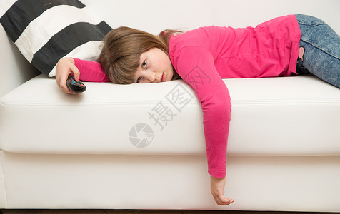 一个无聊的小女孩躺在沙发上看电视在科罗纳检疫期间背景图片
