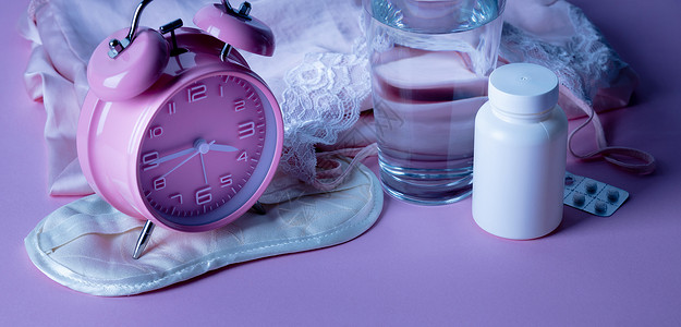 可爱小闹钟粉红色背景的闹钟眼罩药丸和水杯失眠或无睡夜晚的抑郁治疗闹钟药丸和粉红色背景的水杯背景