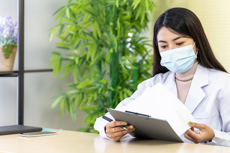 Asian女医生的肖像是戴保护面罩的亚洲女医生坐在院诊所的办公室里在检查耐心之前先阅读文件然后检查耐心背景图片