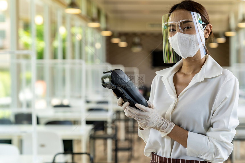 具有吸引力的肖像亚洲女服务员戴面罩和持有信用卡阅读器可以使用室内餐厅背景的无接触支付图片