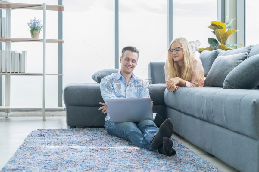 微笑的生意金发白夫妇放松使用电脑笔记本在家里工作和沙发上聊天花时间在隔离冠状概念图片