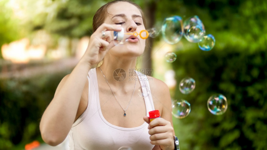 快乐的微笑女人肖像在夏日公园吹着彩色肥皂泡沫快乐的微笑年轻女人肖像在夏日公园吹着彩色肥皂泡沫图片